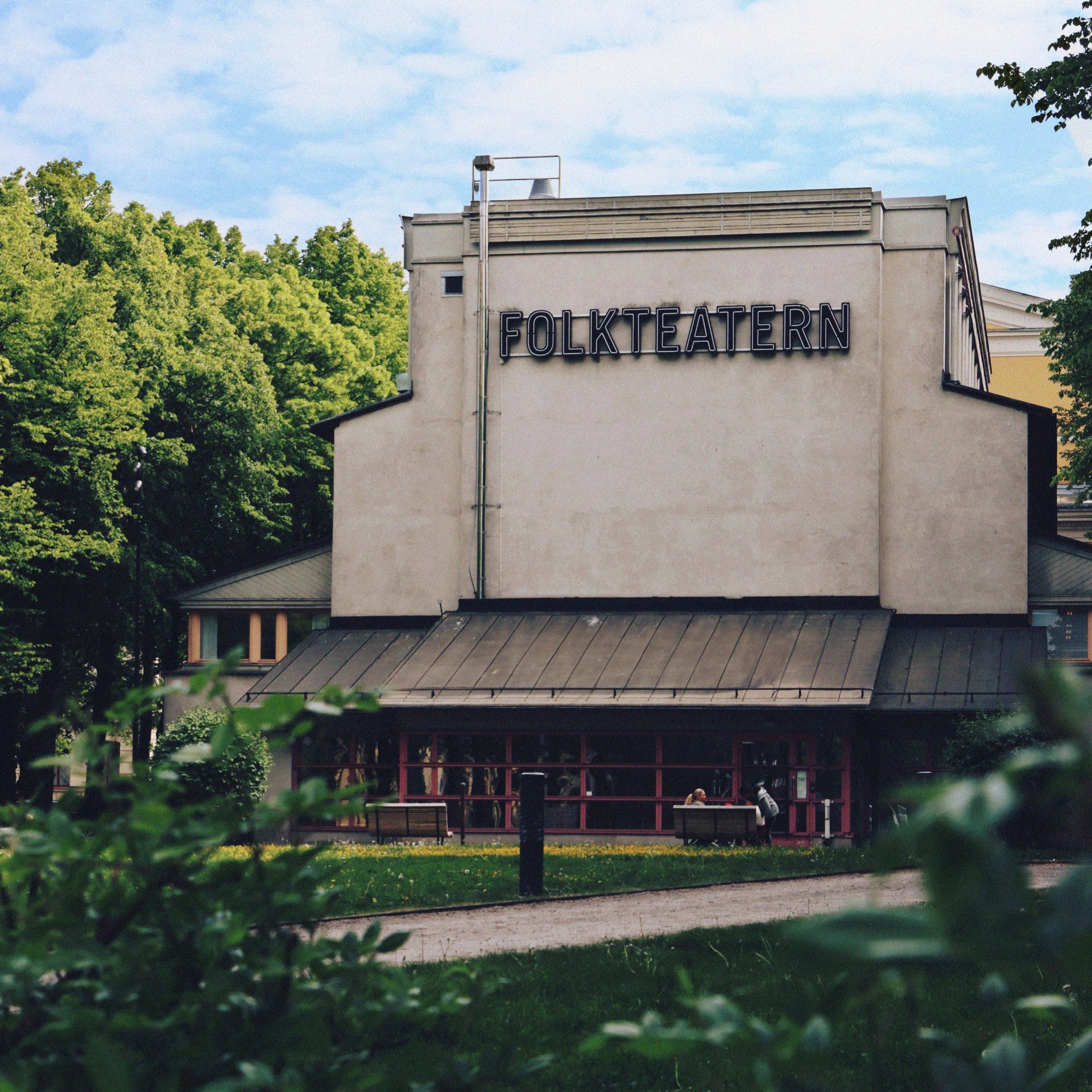 Foto på Folkteatern Gävleborgs teaterhus i Gävle. Huset är inbäddat i grönska från träd, med grusgångar i förgrunden som leder fram till den inglasade entrén